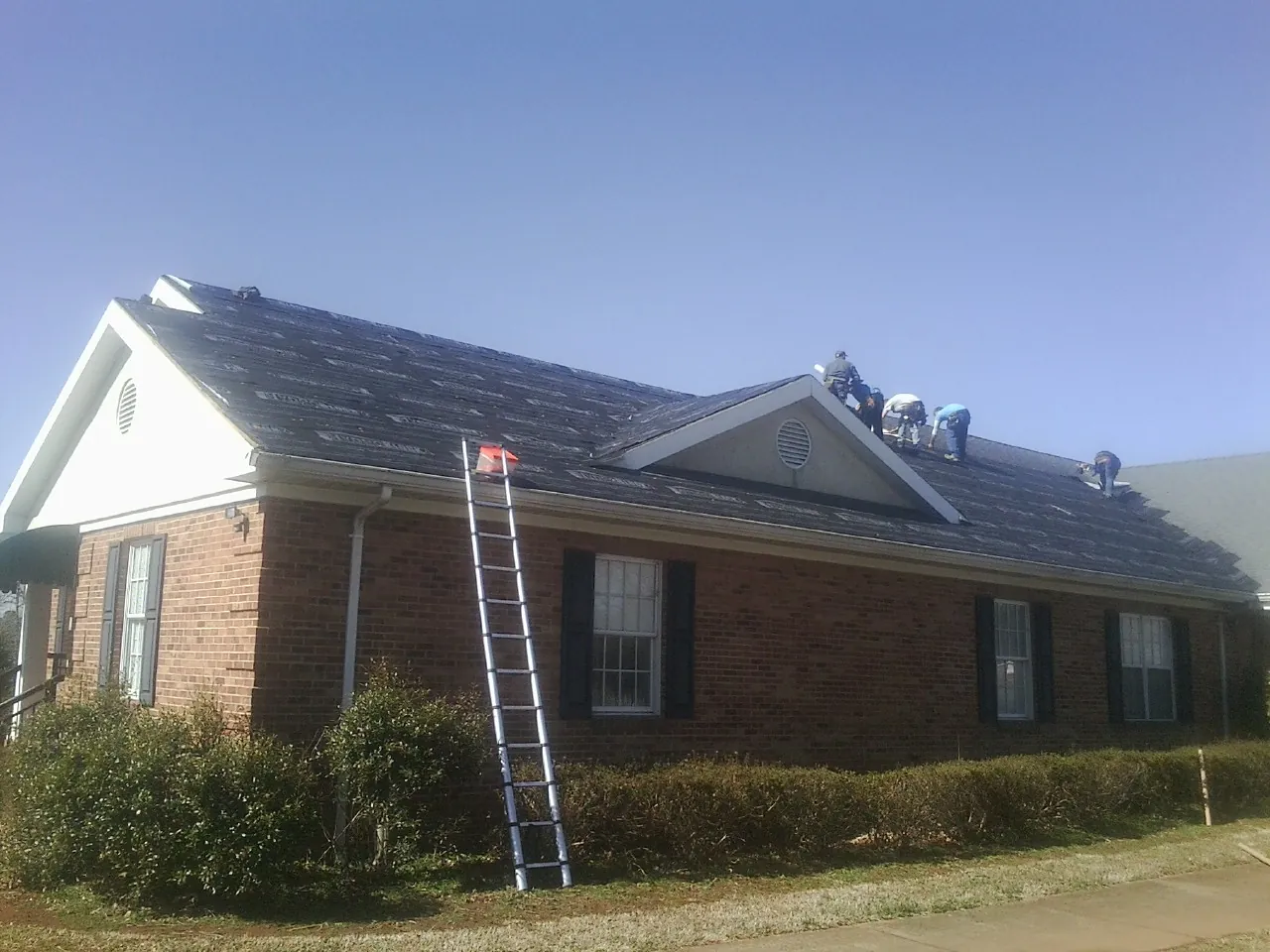 Crew tearing shingles off a brick ranch roof with a ladder set — Metal Roof Repair work by our crew in Bogota
