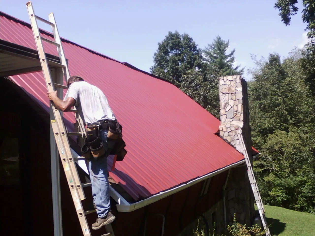 Roofer on a ladder beside a red metal roof and stone chimney — Soffit & Fascia Repair by Roofing Brothers in Bogota